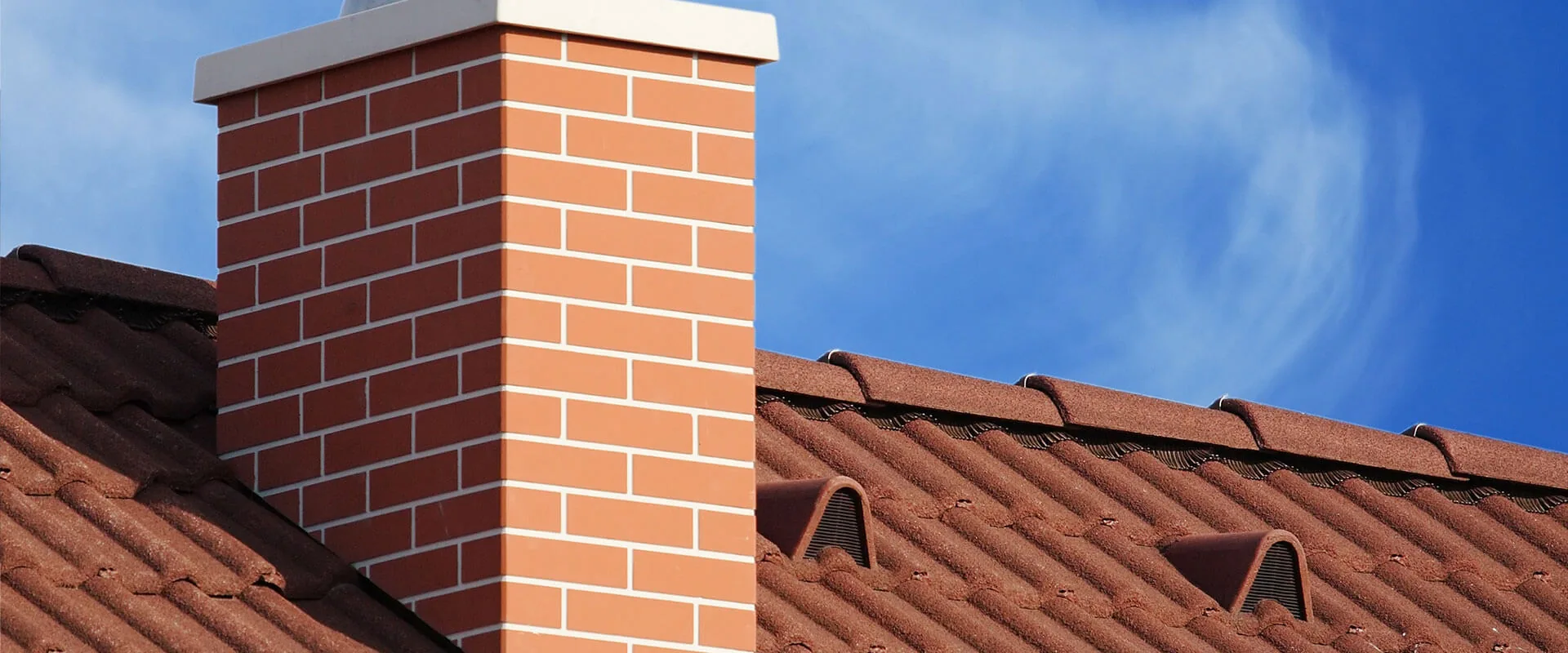 Red brick chimney with metal cap on tiled roof against blue sky.